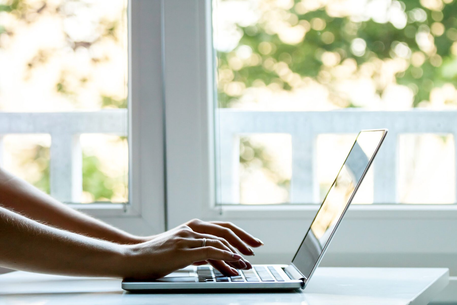 Woman with a ring typing on a laptop with a window and trees in the background.